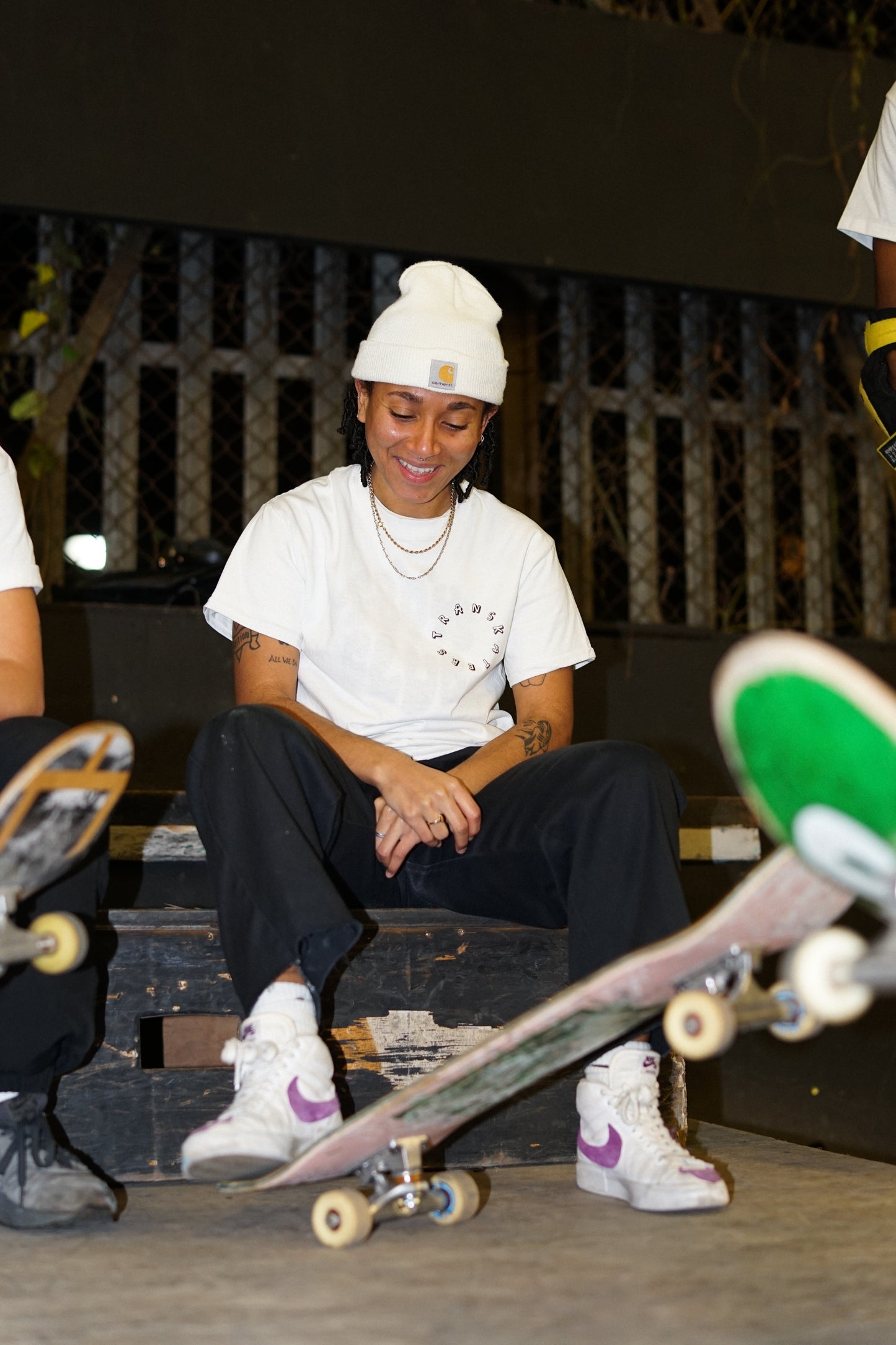 a skateboarder sits on a wooden ledge and smiles and looks at their feet, with one foot on their skateboard 