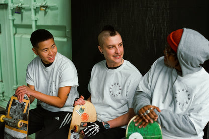 3 skaters wear white Transkaters t-shirts, the person on the left smiling looking at the others, the middle person rocks a small Mohawk, and the person on the right wears rings on their fingers as they rest on the top of their skateboard, a hoodie, hat and glasses