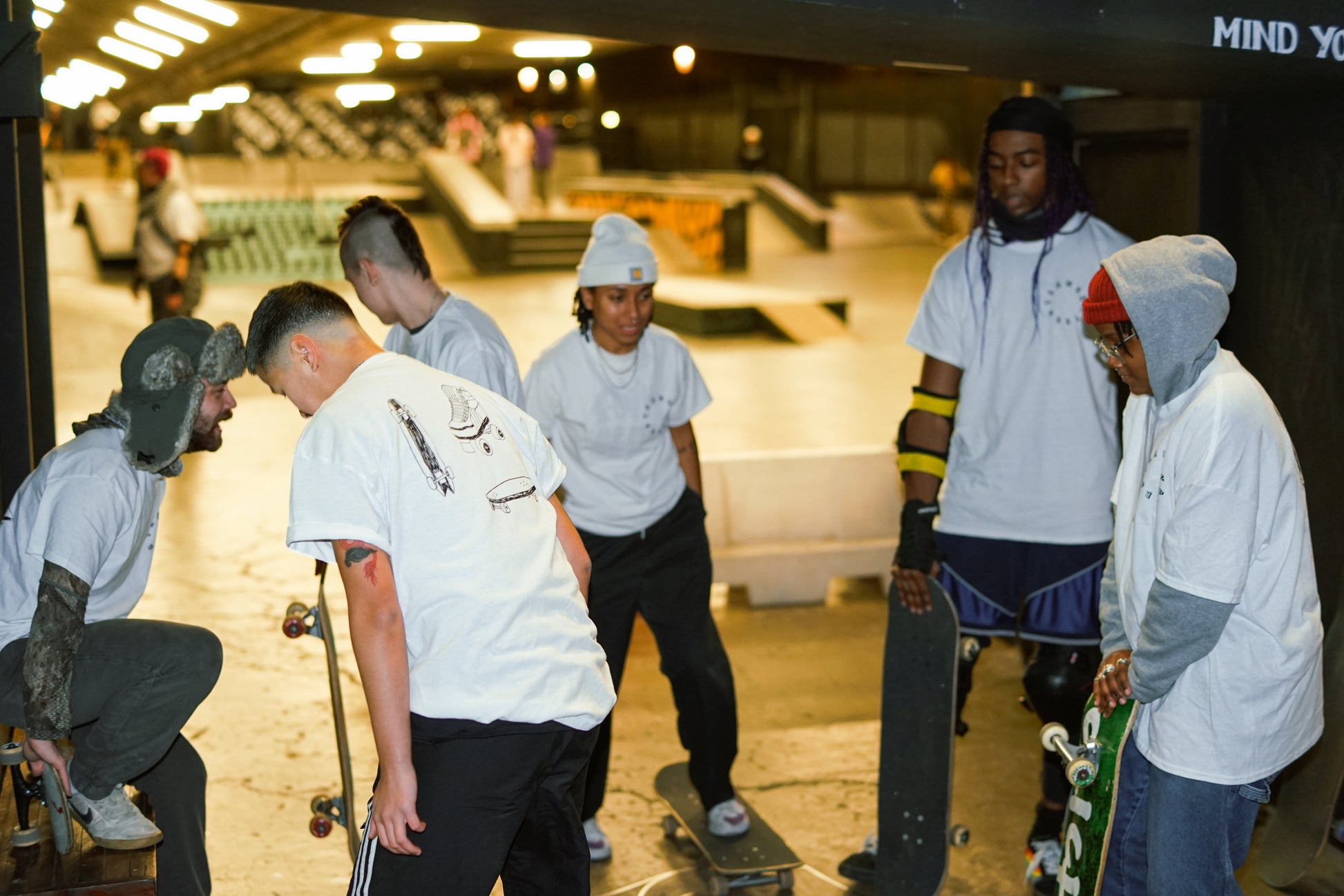 a scene of 6 skaters, all wearing Transkaters white t-shirts, the person on the far left wearing a winter hat and facing the group, the person in the middle standing on the skateboard, and two skaters in the right looking down holding their skateboards 