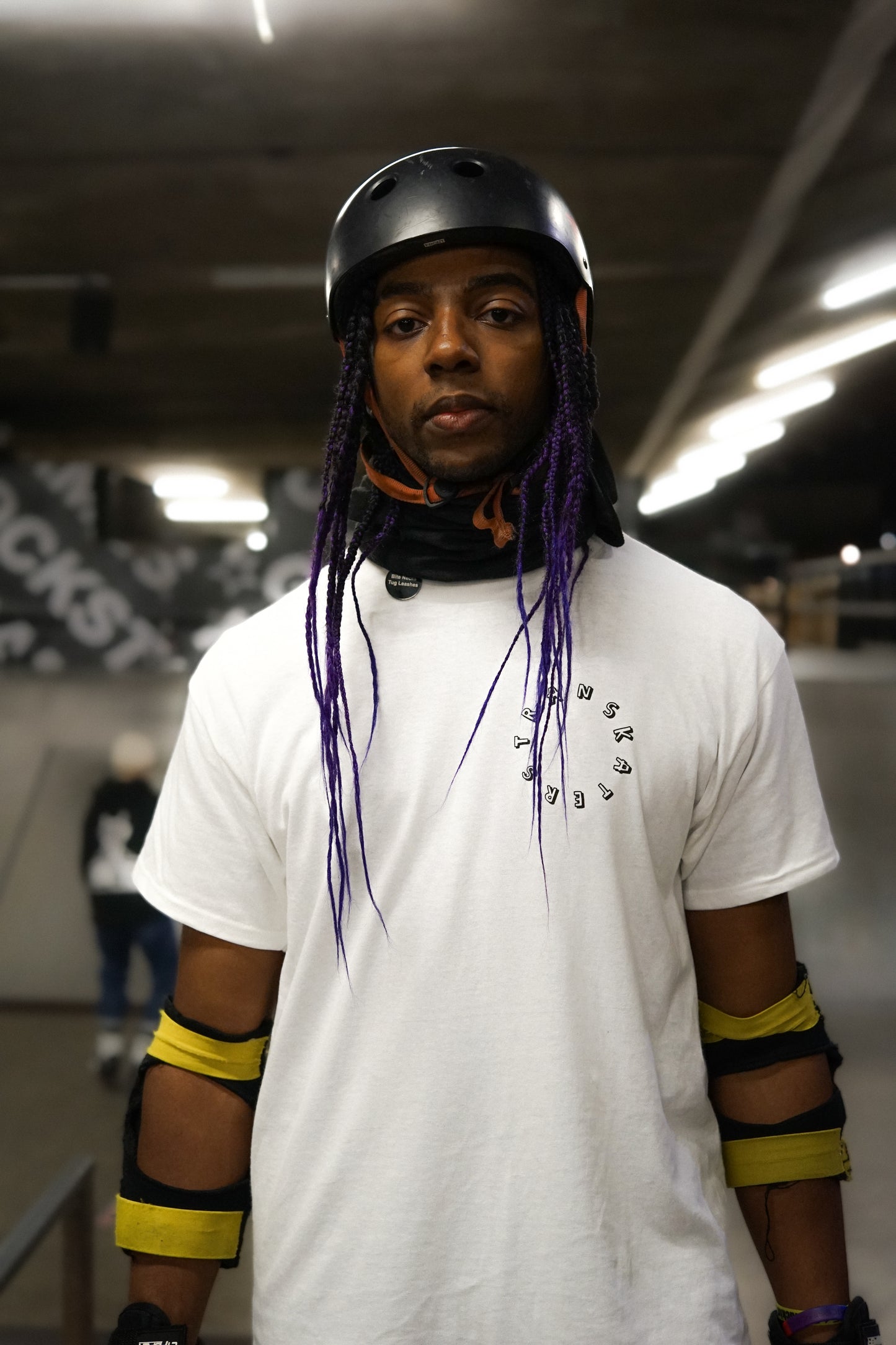 a portrait of a skater with purple braids wearing elbow pads and their white Transkaters t-shirt
